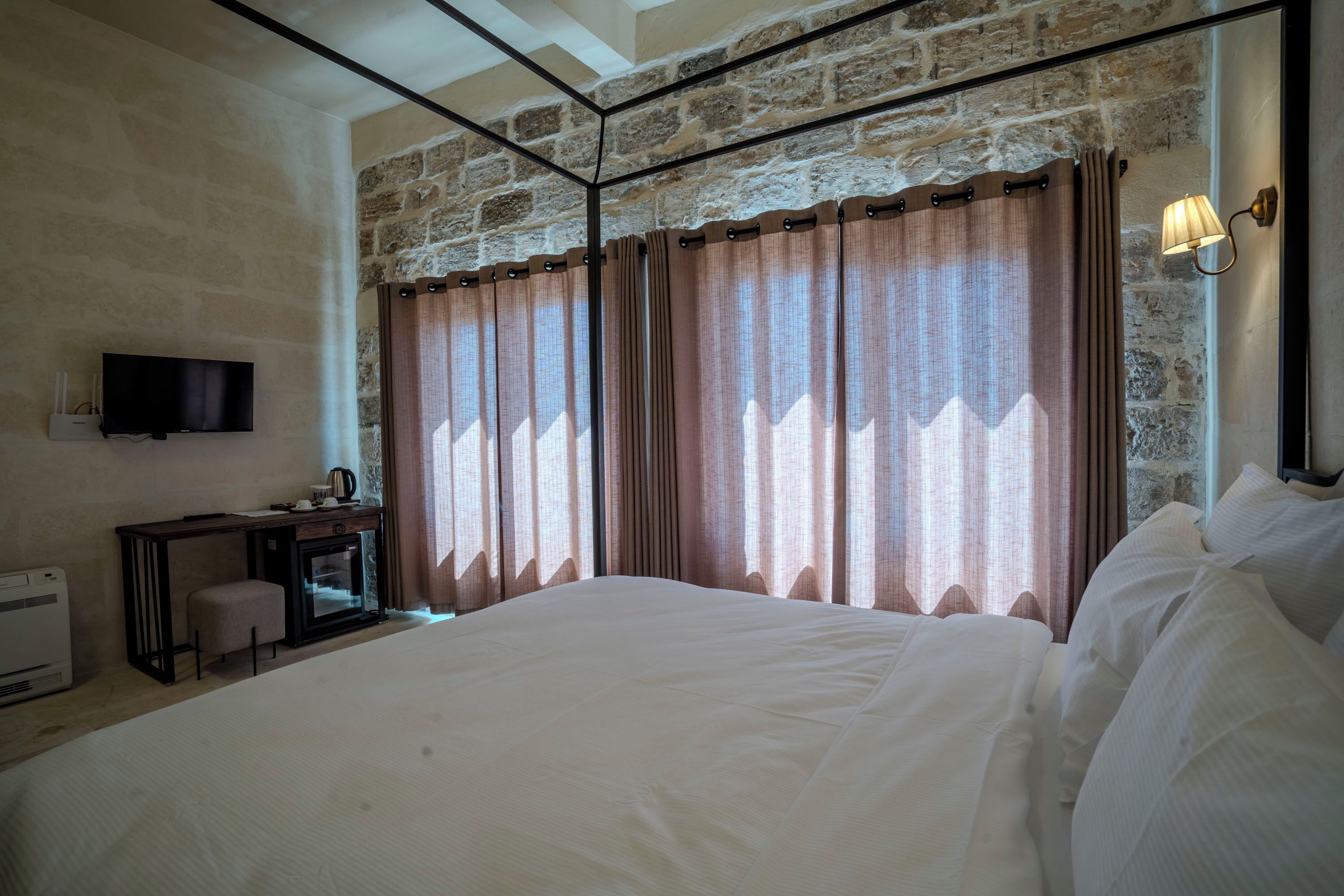 Quarto com cama grande, escrivaninha com cadeira, televisão e parede de pedra.