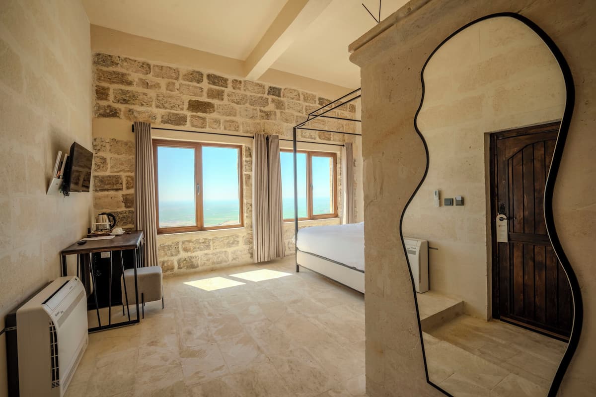 A room with stone walls, a bed, a desk with a chair, and a view of the sea through large windows.