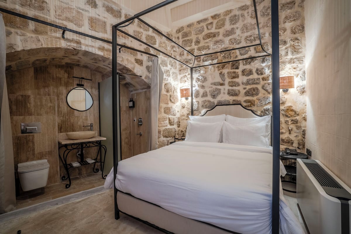 A bedroom with a stone wall, a bed with white linens, a bedside table, and a round mirror.