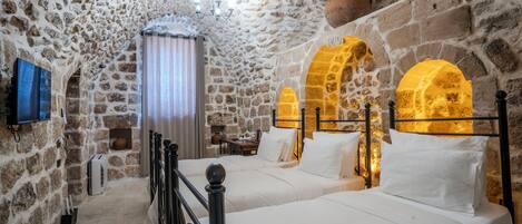 A stone-walled bedroom with two beds, a television, and a window with curtains.