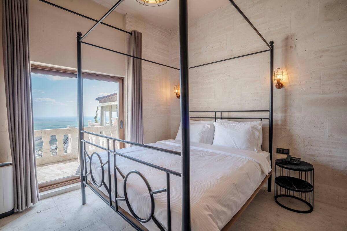 A four-poster bed with a view of the sea, a round side table, and a wall-mounted light fixture.