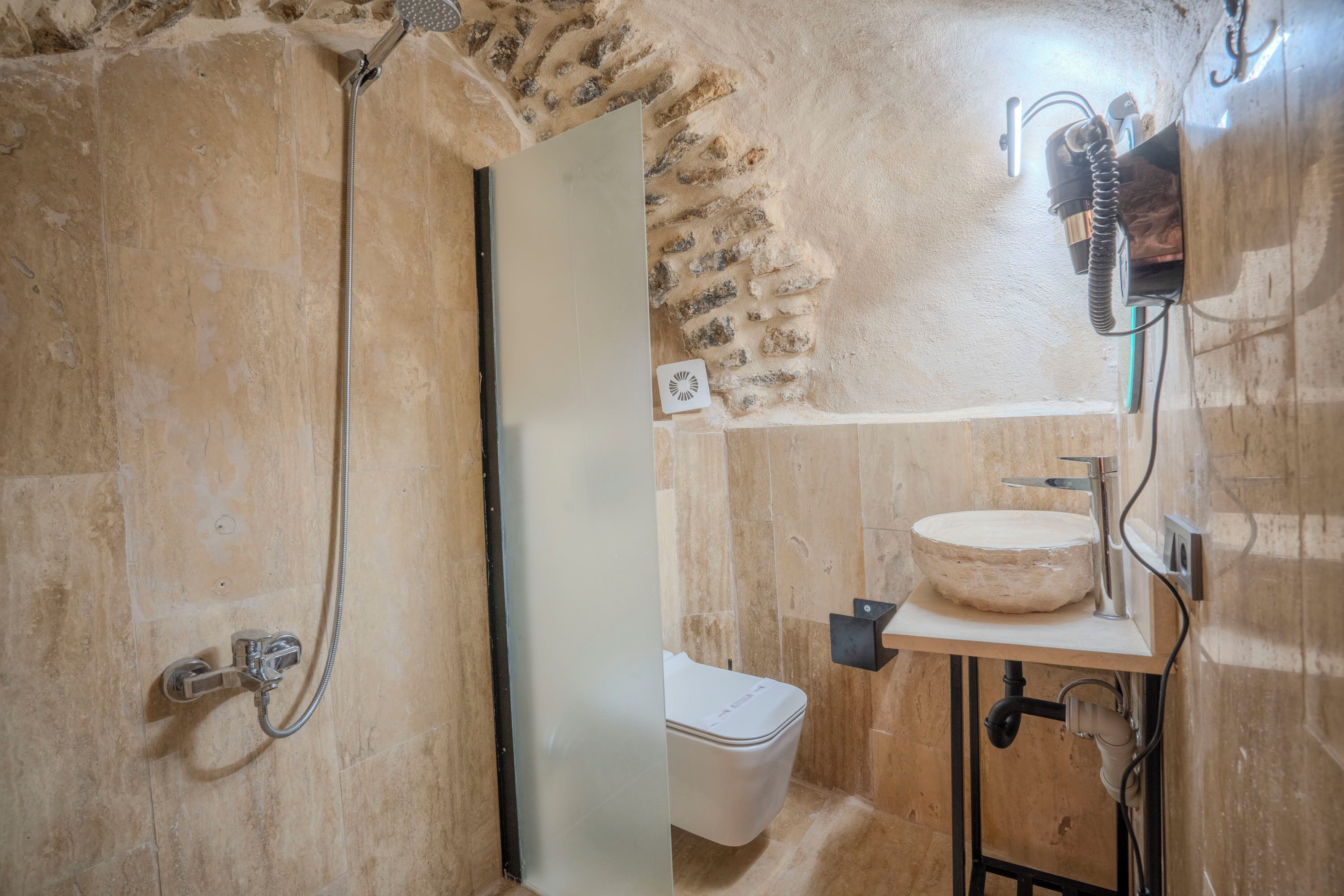 A bathroom with a stone wall, a white toilet, a sink with a round basin, and a shower with a handheld showerhead.