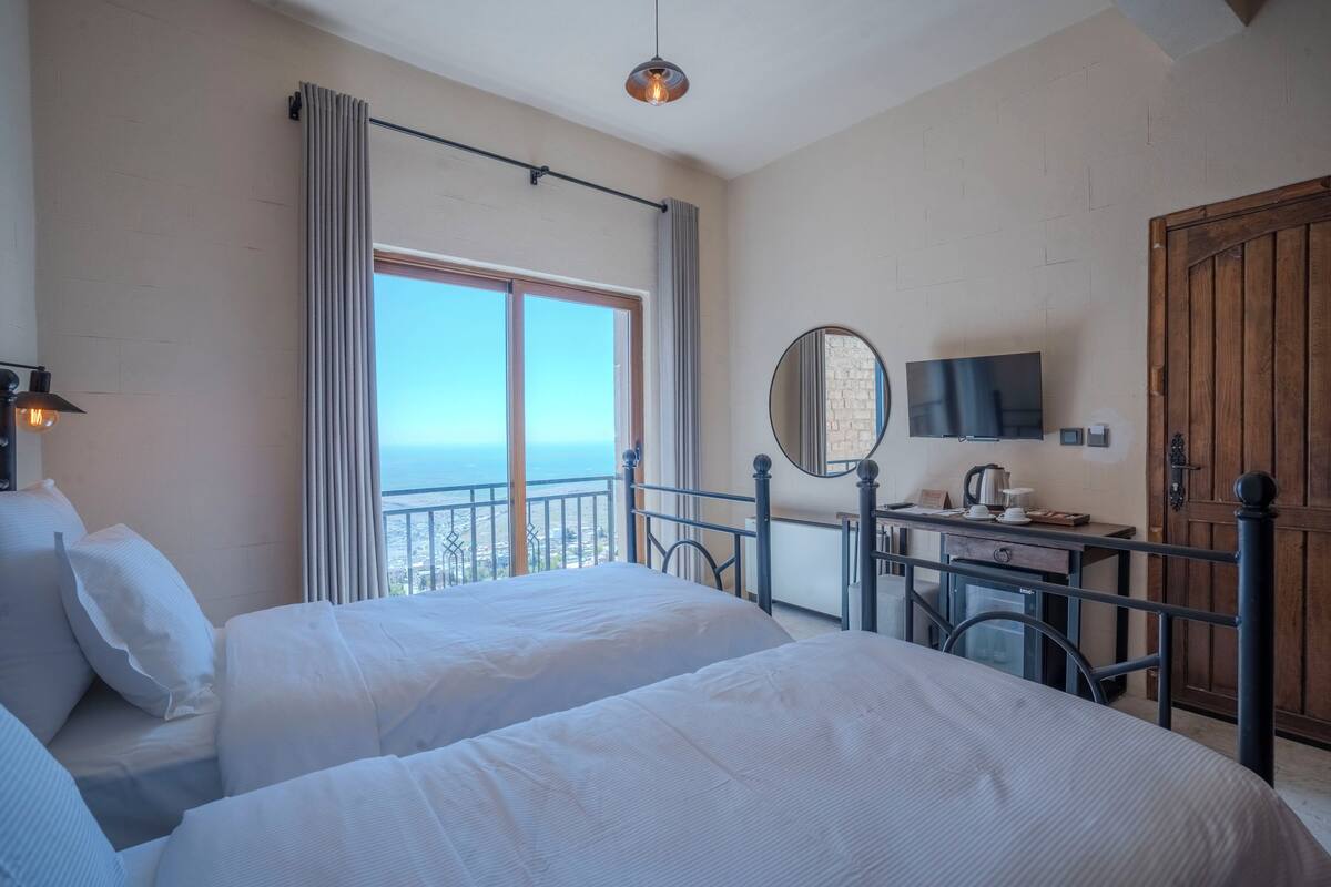 A hotel room with two beds, a desk with a mirror, a television, and a view of the sea.