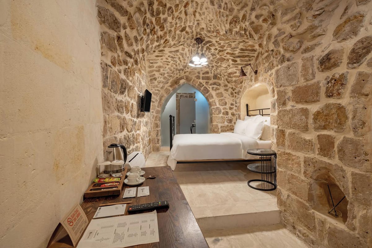 A stone-walled room with a bed, a small table with items, and a doorway leading to another room.