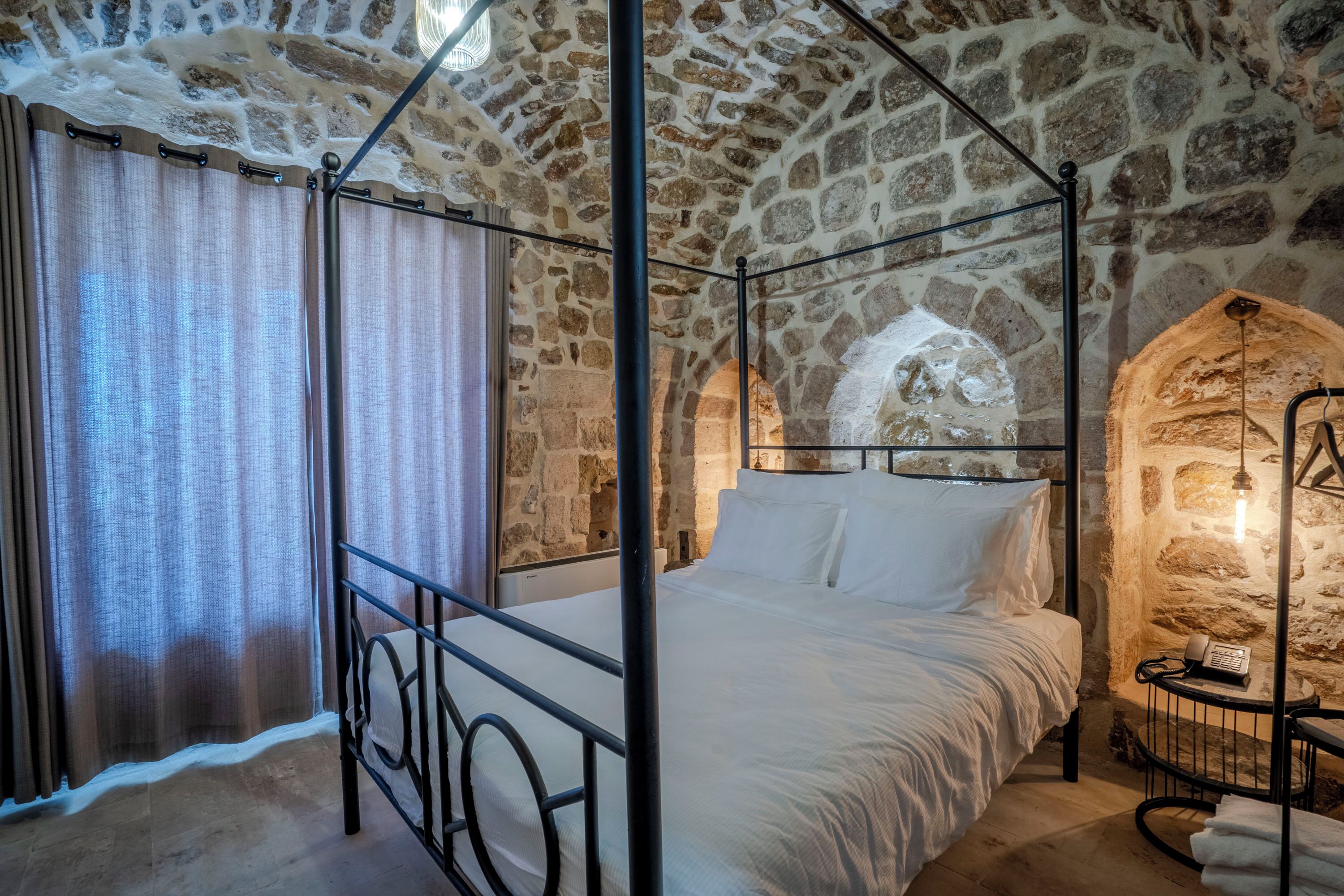 A four-poster bed in a stone-walled room with a stone archway and a small table with a telephone.