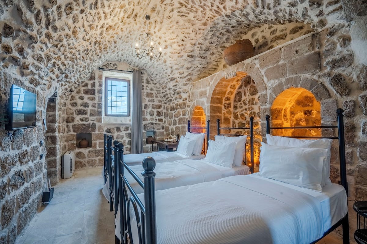 A stone-walled bedroom with two beds, a window, and a chandelier.