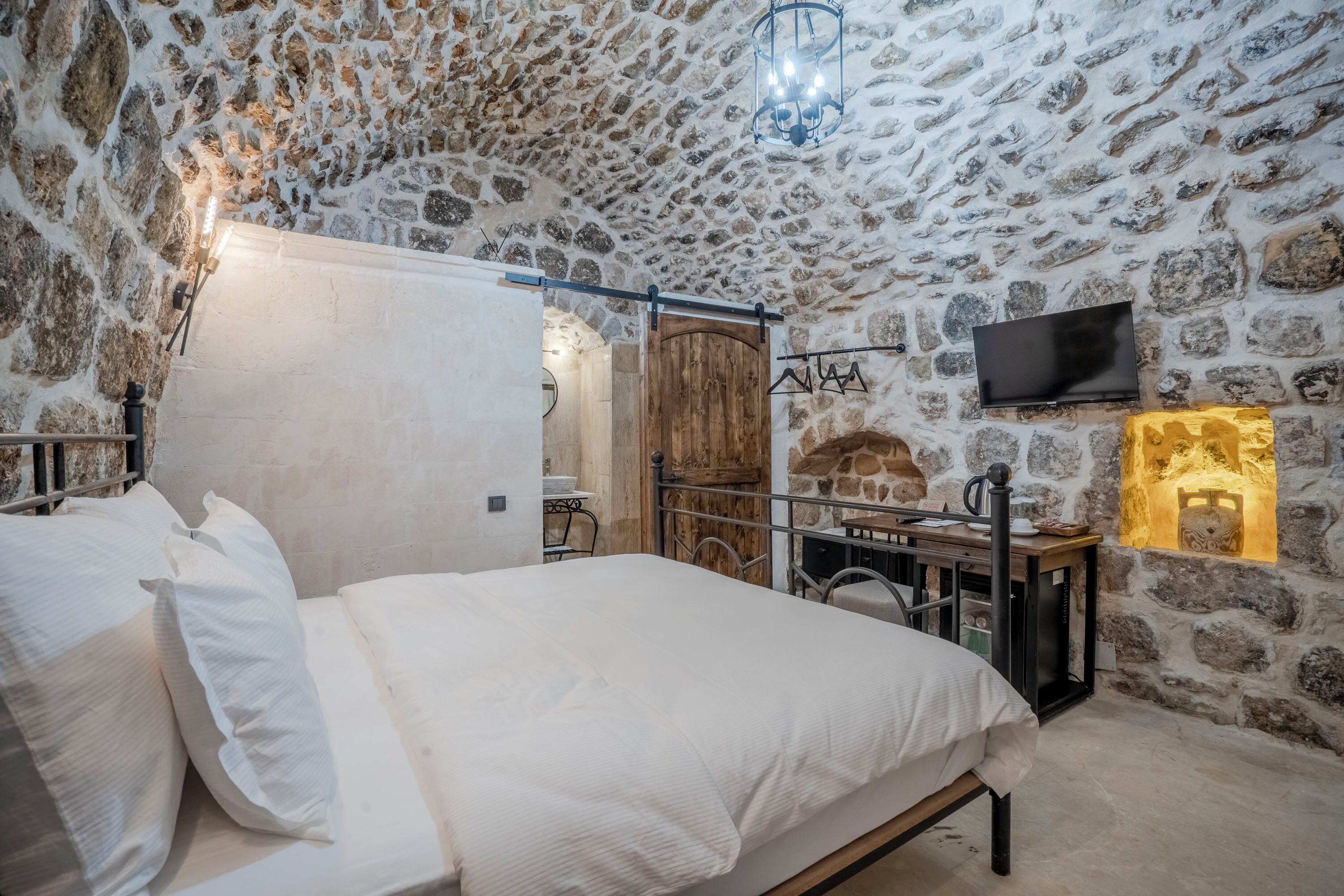 A bedroom with a stone wall, a bed with white bedding, a wooden wardrobe, a television mounted on the wall, and a small table with a lamp.