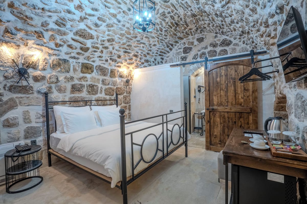 A stone-walled bedroom with a bed, a wooden wardrobe, and a small table with a kettle and cups.