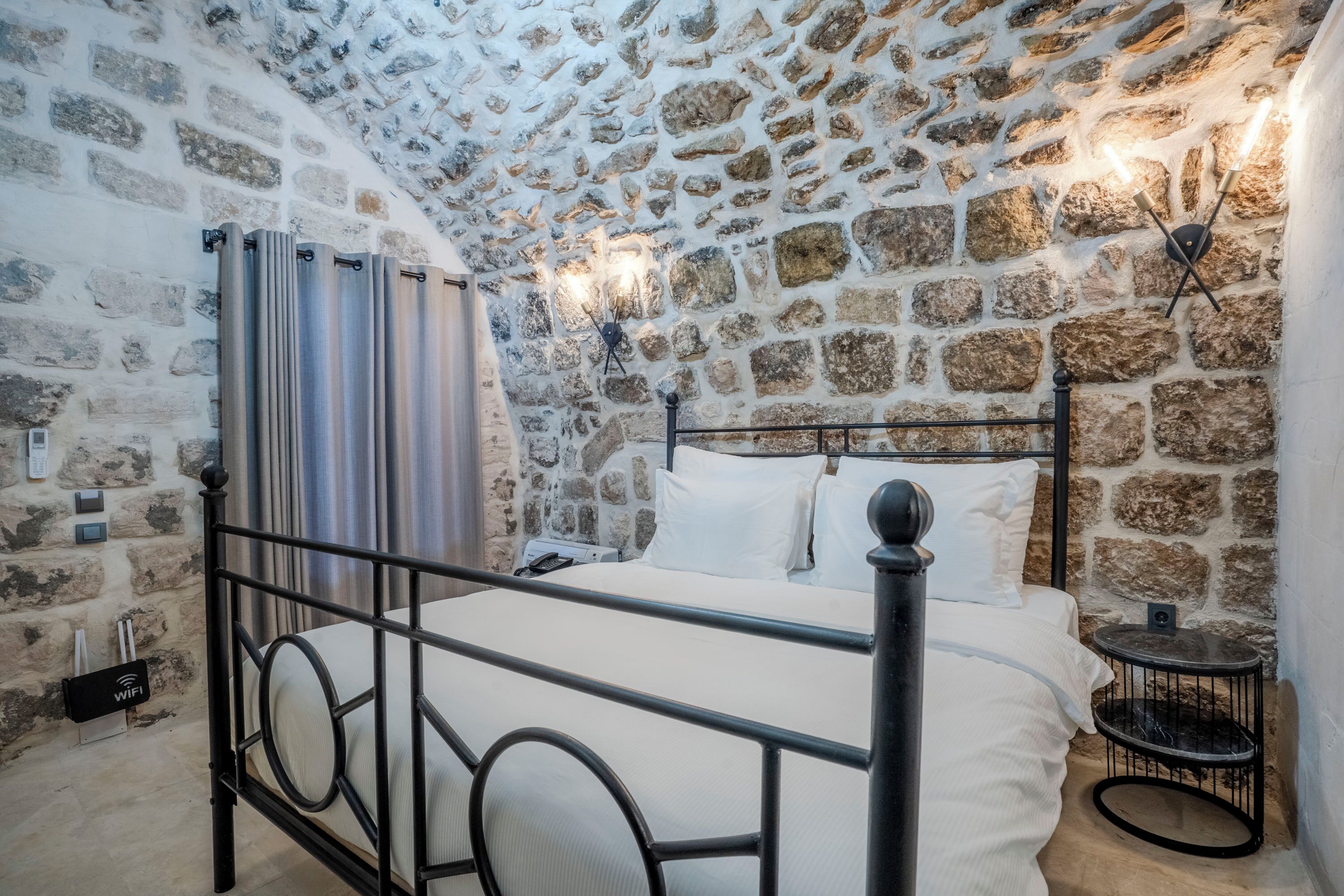 Un dormitorio con pared de piedra, estructura metálica para la cama, ropa de cama blanca, una mesita de noche pequeña y un aplique de pared.