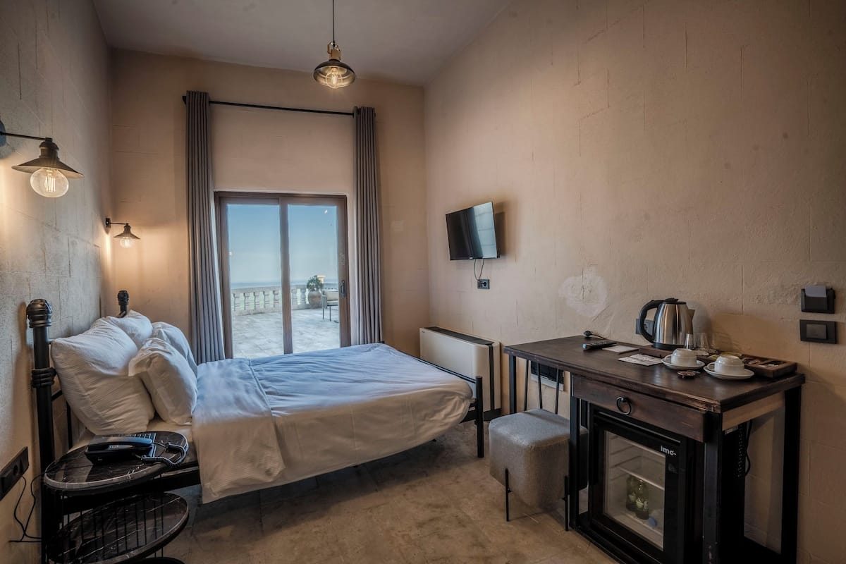 A hotel room with a bed, a desk with a kettle, a TV, and a window with curtains.