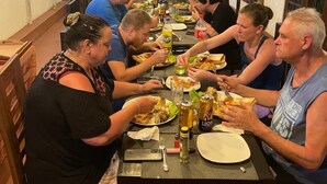 Een groep mensen dineert aan een lange tafel in een binnenruimte, met hangende lampen en decoratieve details.