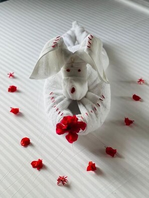 A white, plush, stuffed bunny toy with red stitching and a red flower attached, placed on a white, striped surface with scattered red petals. - Villa Acadia  (Dharga Town)