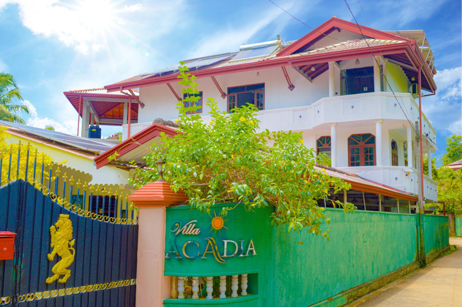 A two-story building with a red roof and white walls, surrounded by a green fence with a lion emblem and the name "Villa Acadia".