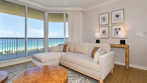 A living room with a large window overlooking a beach, a beige sectional sofa, a round coffee table, and a console table with lamps. - Stunning beachfront condo w/ relaxing amenities in the heart of Destin! 🐚🏖🐟 (Destin)