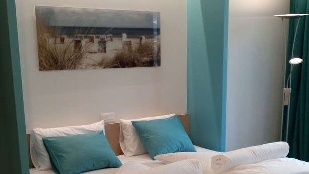 A neatly made bed with white linens and blue pillows, a painting of a beach scene on the wall, and teal-colored furniture. - Your bed by the sea (Koksijde)