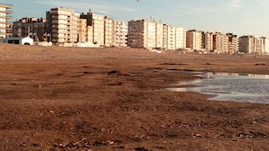 A sandy beach with footprints and a bird flying in the sky. In the background, there are multi-story buildings. - Your bed by the sea (Koksijde)