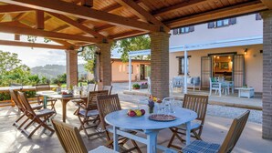 An outdoor patio with wooden tables and chairs, a covered area with a wooden ceiling, and a view of a building in the background. - Casale Partemio 8 Marche Holiday Villas (Cartoceto)
