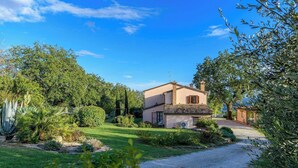A two-story house with a tiled roof, surrounded by a well-maintained garden with various plants and a gravel driveway. - Casale Partemio 8 Marche Holiday Villas (Cartoceto)