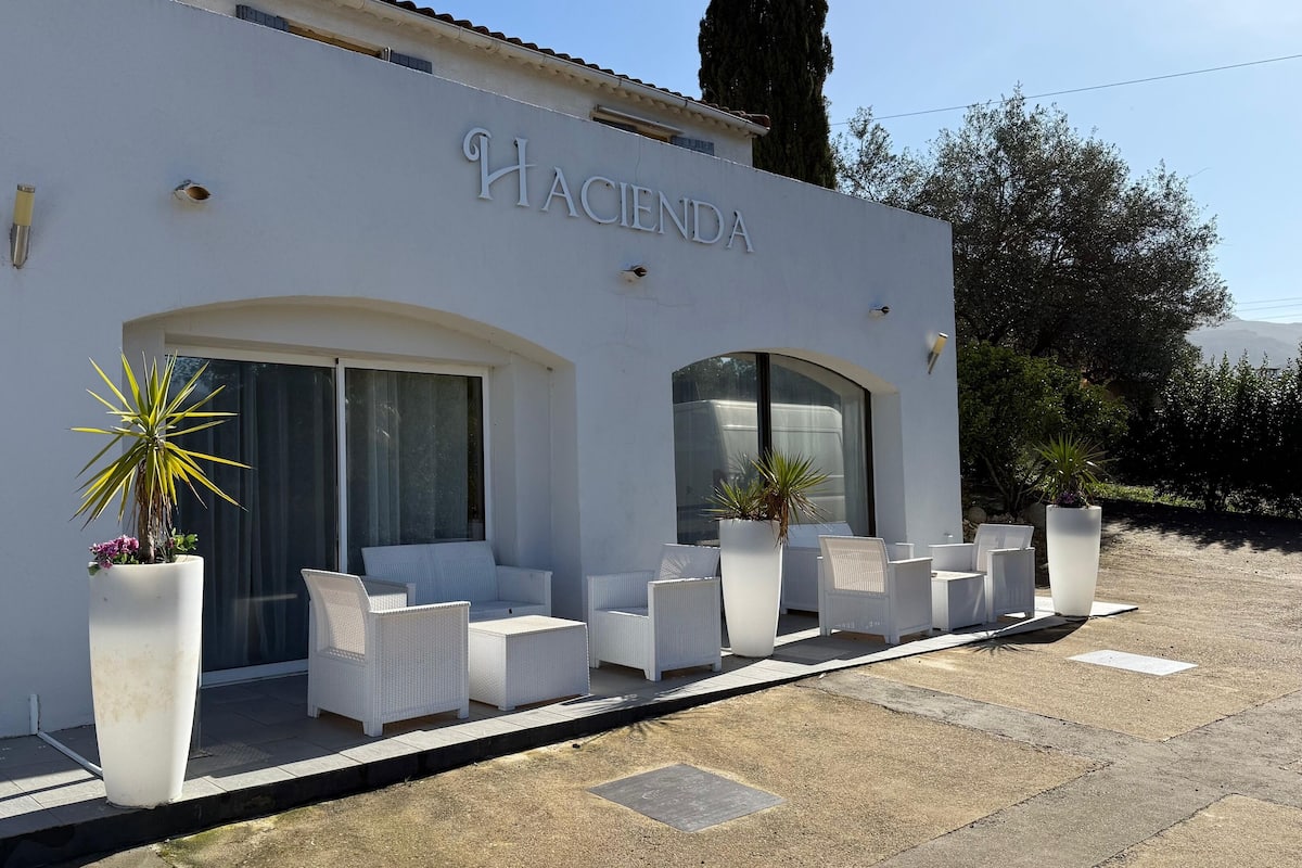 A building with the name "Hacienda" on the facade, featuring a patio with white wicker furniture and large potted plants.