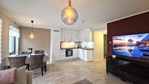 A modern living room with a dining area, a kitchen, and a large TV screen displaying a sunset view.