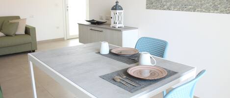 A dining area with a table set for two, a green sofa, a small kitchenette, and a painting on the wall.
