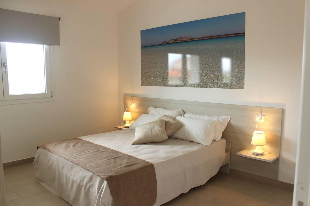 A bedroom with a bed, bedside lamps, a window with a blind, and a painting of a beach scene.