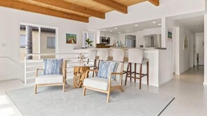 Un salon moderne avec un plafond en bois, un coin repas avec des chaises blanches et une cuisine à l’arrière-plan.