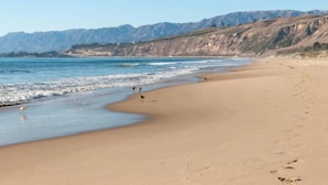 Une plage à l’eau d’un bleu limpide, avec un rivage sablonneux et des montagnes en arrière-plan.