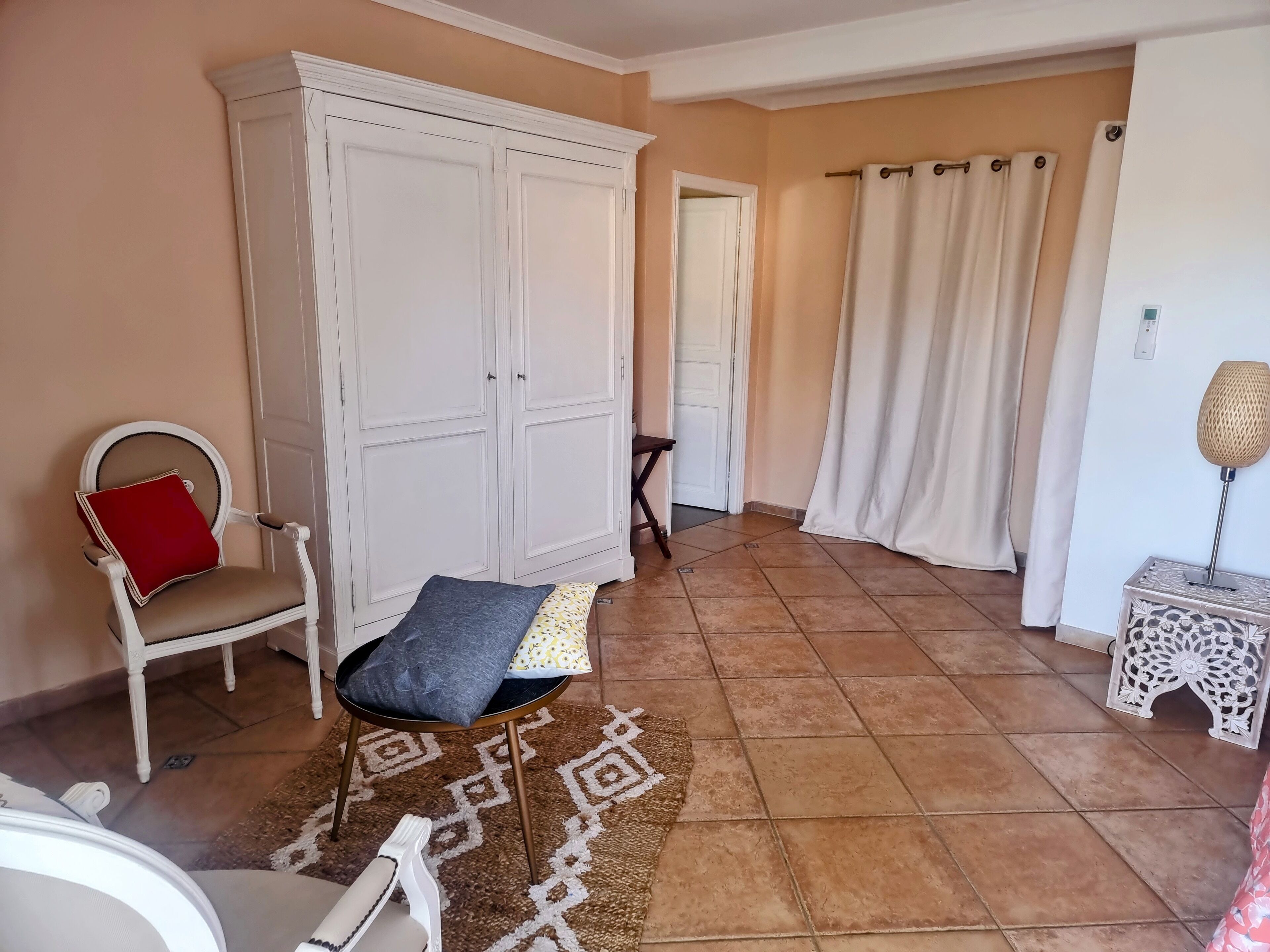 A room with a white wardrobe, a chair with a red cushion, a patterned rug, and a small table with a lamp.