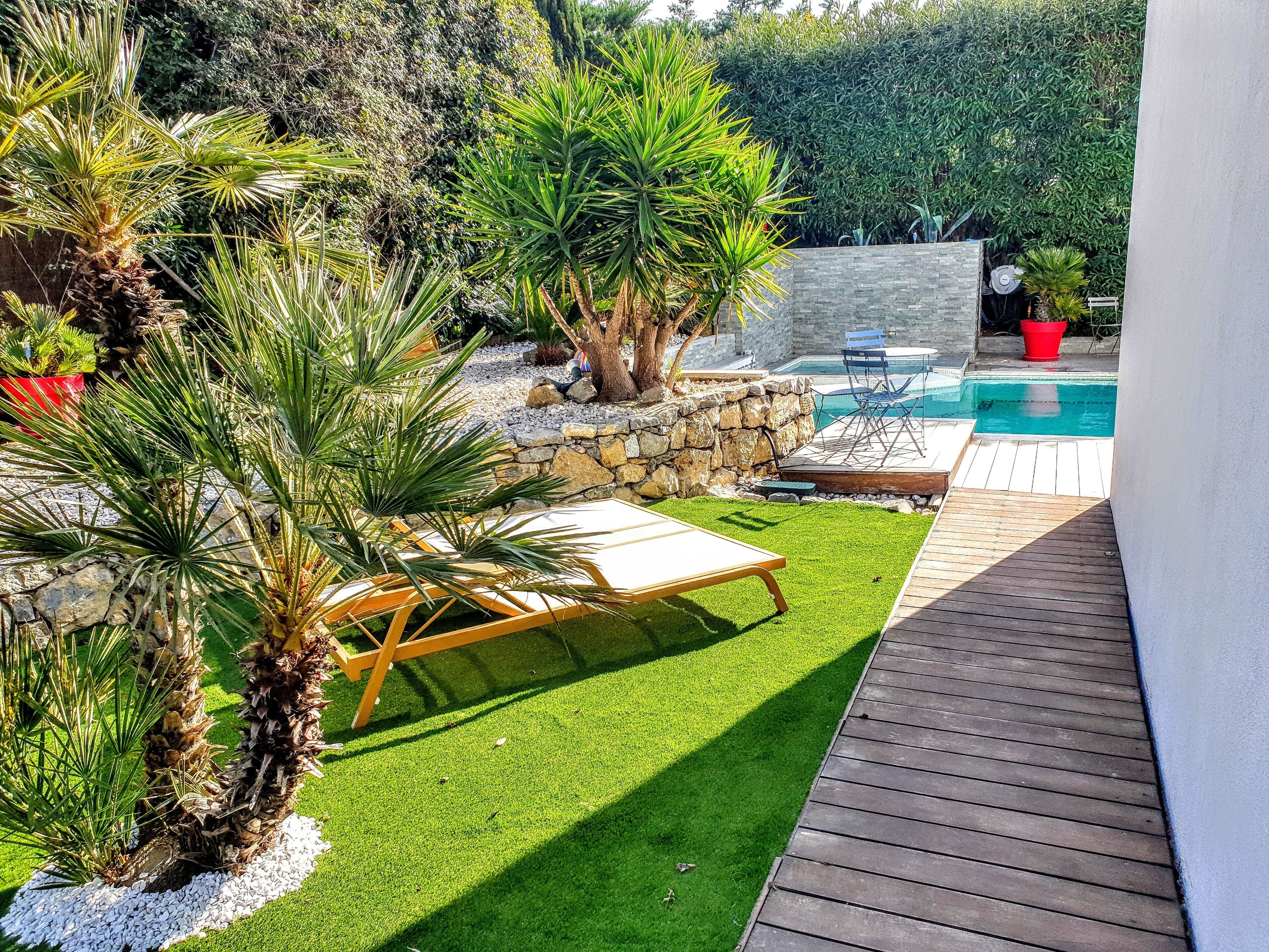 Un patio trasero con piscina, césped artificial, palmeras y un muro de piedra.