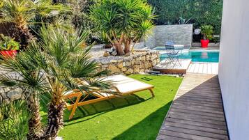 Un jardin avec une piscine, du gazon synthétique, des palmiers et un mur en pierre.