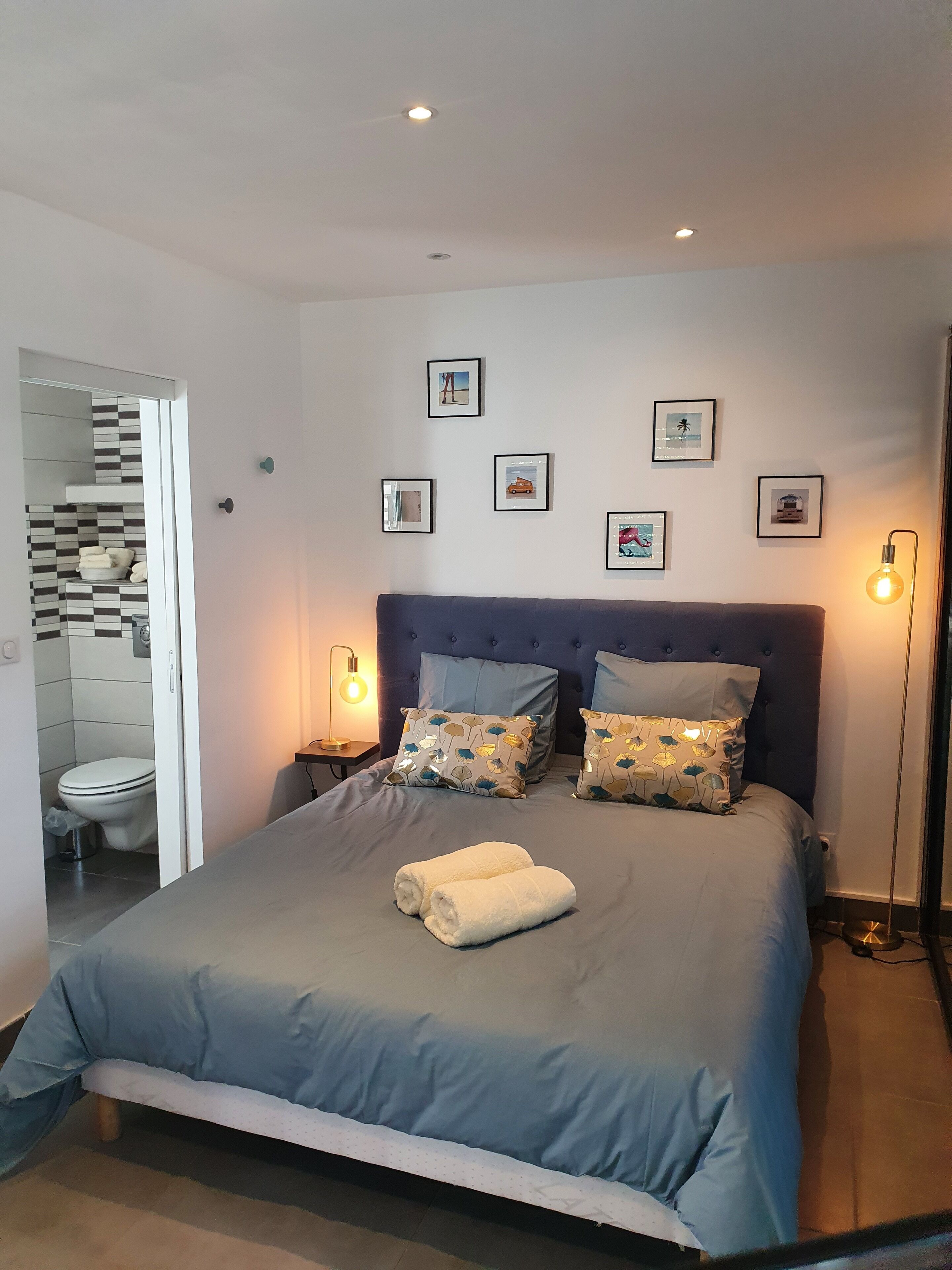 A bedroom with a bed, two pillows, a bedside table, a floor lamp, and a wall with framed pictures.