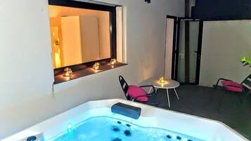 A hot tub with a window above it, a small table with a candle, and a chair.