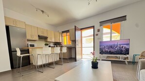 Une cuisine moderne équipée d’un réfrigérateur, d’un four et d’un four à micro-ondes. On y trouve un coin repas avec une table blanche et des tabourets de bar. Un téléviseur est fixé au mur, et une fenêtre apporte de la lumière naturelle.