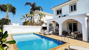 Un espace piscine avec des chaises longues, un bâtiment blanc doté d’un toit en tuiles et des palmiers.