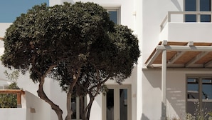 A modern white building with a staircase leading to a door, a tree in the foreground, and a clear blue sky. - Akria Seaside Suites (Paros)