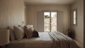 A bedroom with a large bed, a bedside table, a lamp, and a view through a glass door to an outdoor area. - Akria Seaside Suites (Paros)