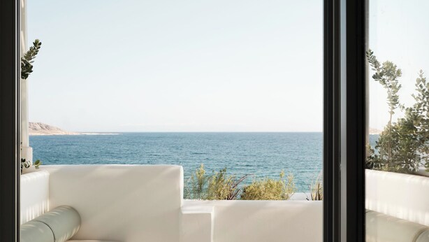 A balcony with a white sofa, a white table with a book and a bowl, and a view of the sea. - Akria Seaside Suites (Paros)