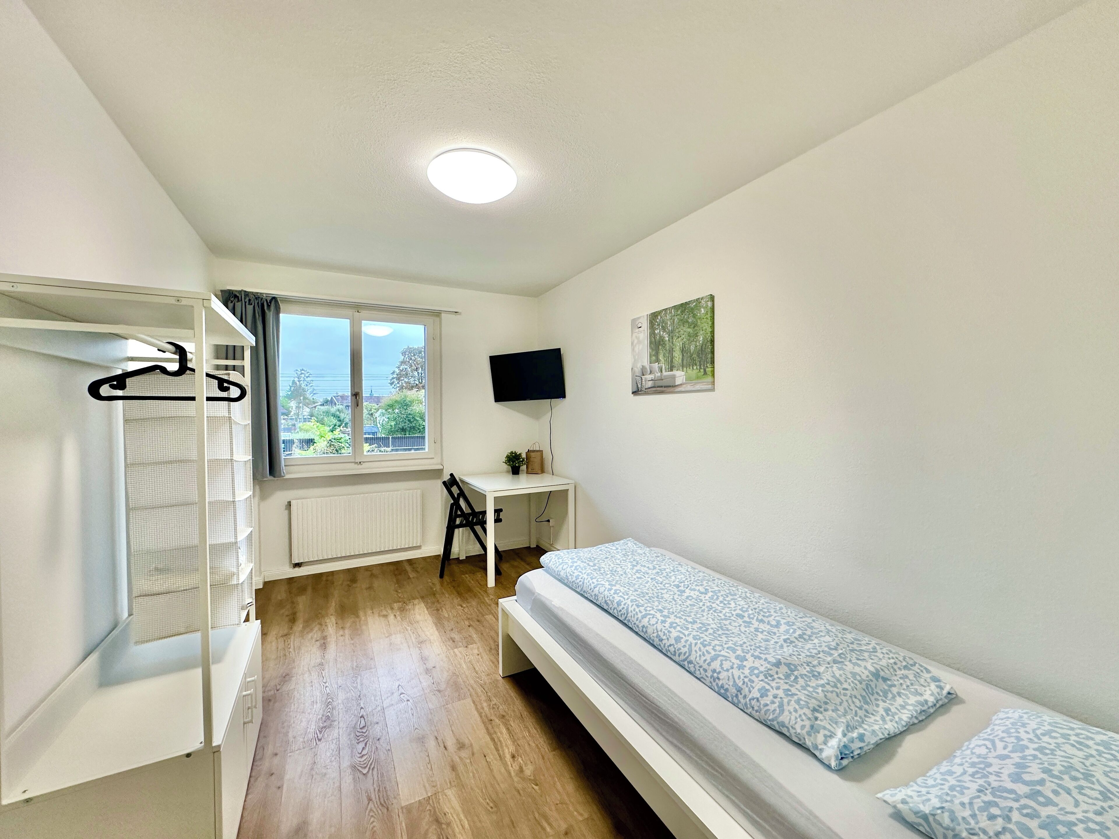 A bedroom with a bunk bed, a desk, a chair, a TV, and a window with a view of greenery.