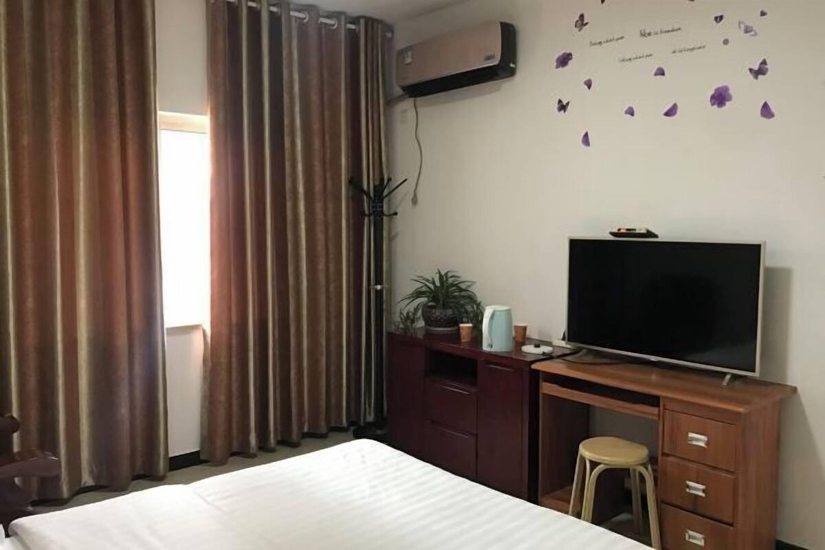 A hotel room with a bed, a desk with a TV, a chair, a plant, and a window with curtains.