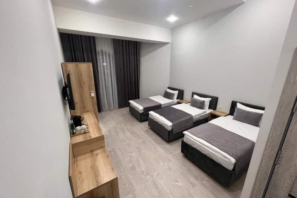 A modern hotel room with three beds, a wooden desk, and a large window with curtains.
