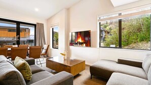 A modern living room with a fireplace, comfortable seating, and large windows offering a view of the outdoors. - Grand Alpine Villa - 4 bedrooms (Queenstown)