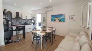 Une cuisine moderne avec des armoires blanches, un comptoir noir et une hotte en acier inoxydable. On y trouve un coin repas avec une table ronde et quatre chaises. La pièce est dotée d'un plancher en bois et d'un canapé beige.