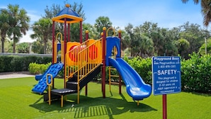 Un parque infantil con toboganes y estructuras para escalar, rodeado de vegetación y un cartel de seguridad.
