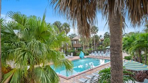 Zona de piscina con sillones de descanso y sombrillas, rodeada de palmeras y un edificio con balcones.