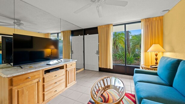 A modern living room with a large TV, wooden cabinets, a blue sofa, and a view of palm trees through the sliding glass doors. - Park Thrills + Poolside Vibes = Vacation Goals! (Kissimmee)