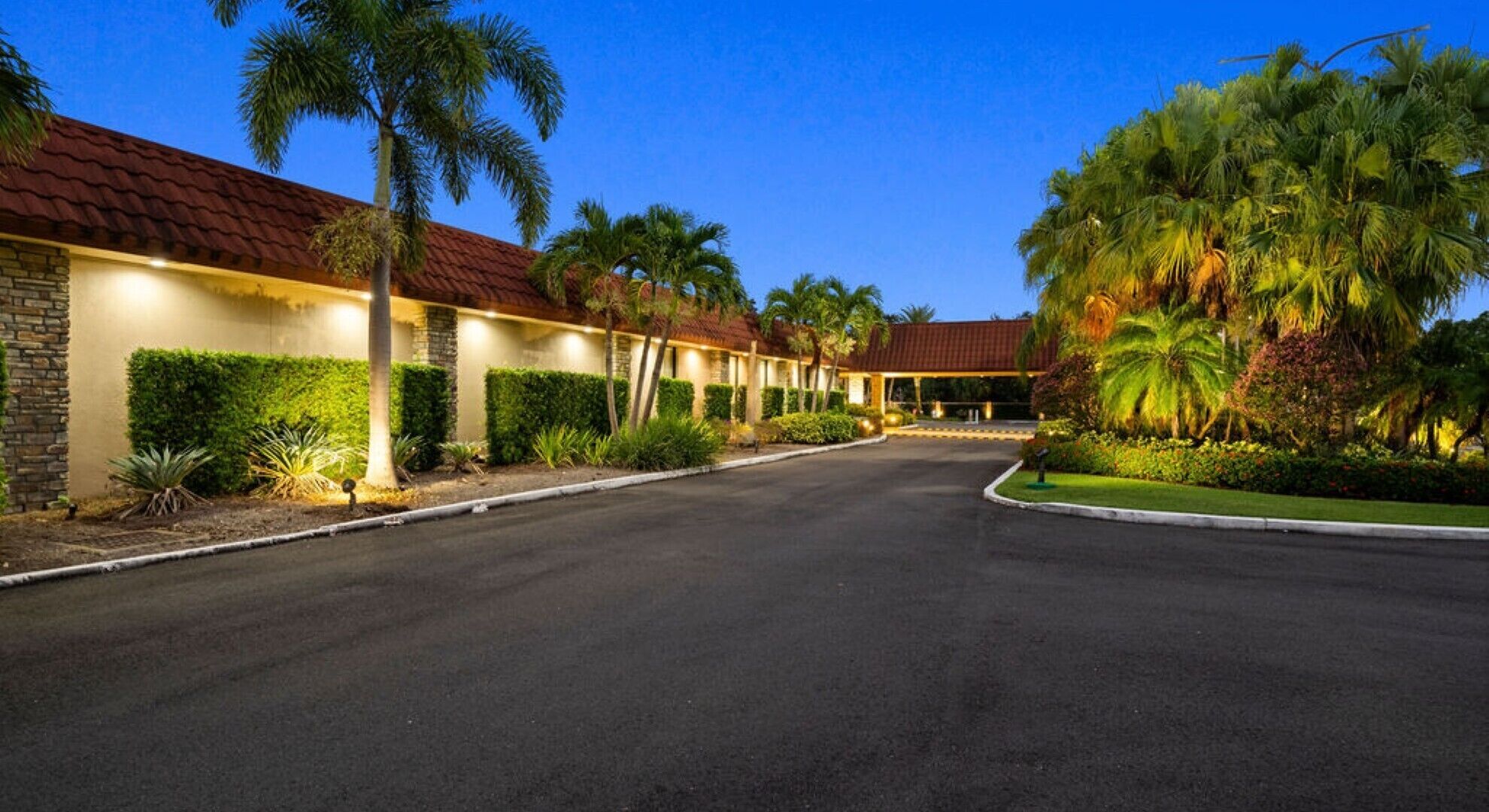 Un hotel con una hilera de habitaciones, un camino de entrada pavimentado y jardines bien cuidados con palmeras y arbustos.