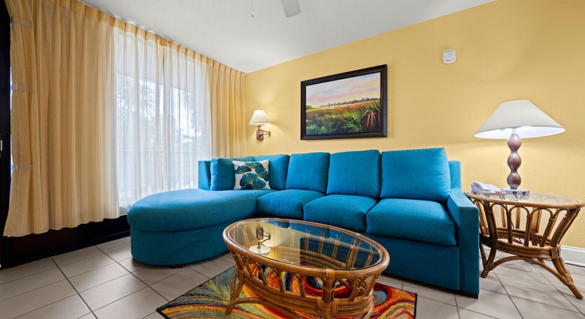 A living room with a blue sofa, a glass coffee table, a wicker side table, a lamp, and a painting on the wall.