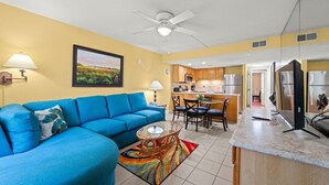 A living room with a blue sofa, a dining area with a table and chairs, a kitchen with wooden cabinets and appliances, and a ceiling fan. - Park-Hopping Made Easy | 2BR Retreat Near Disney & Universal (Kissimmee)