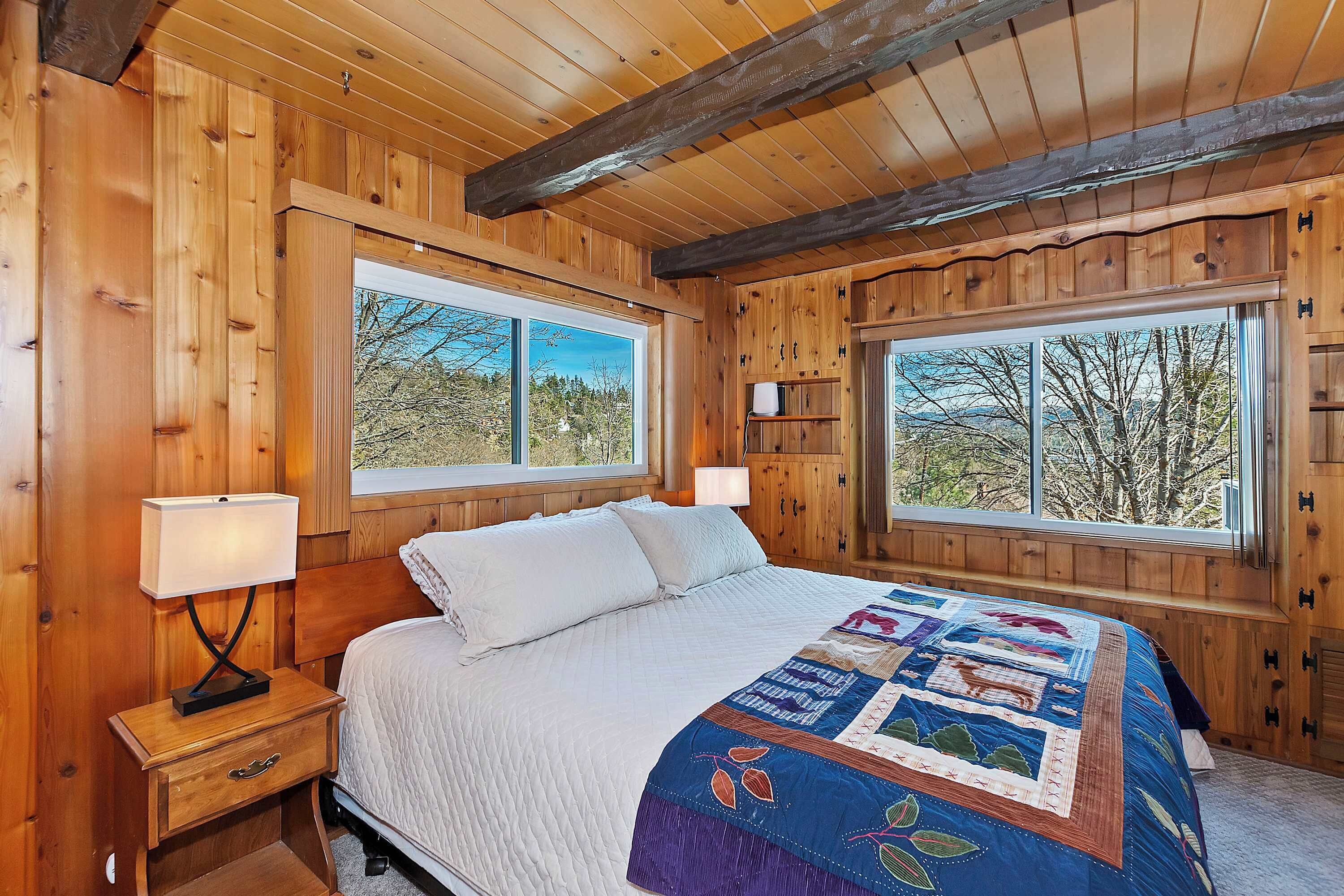 Un dormitorio de cabaña de madera con una cama grande, dos ventanas, una mesita de noche y un colcha.